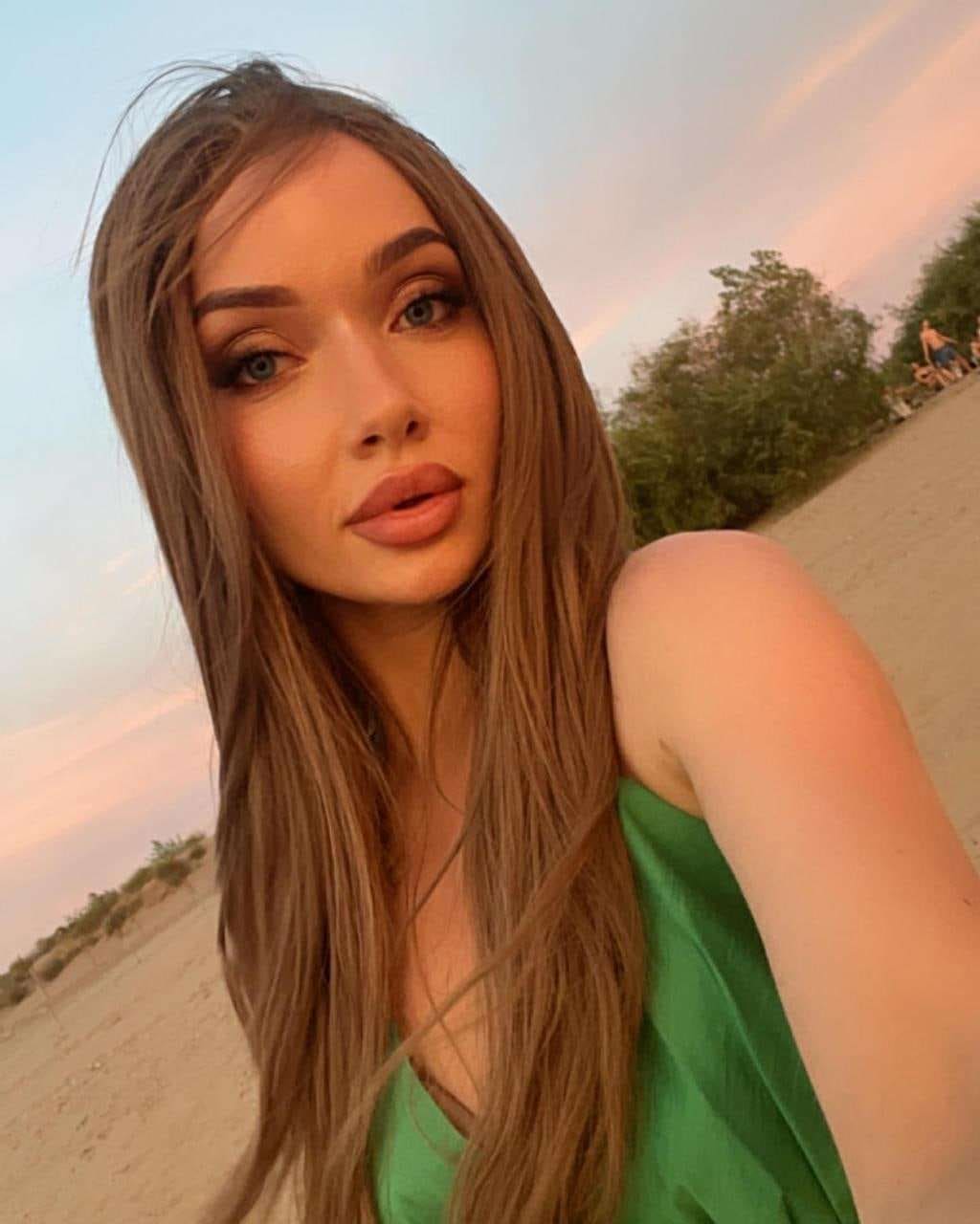 Agatha takes a selfie on a sandy beach at dusk, wearing a sleeveless green V-neck top with bare shoulders visible against a pink sky.