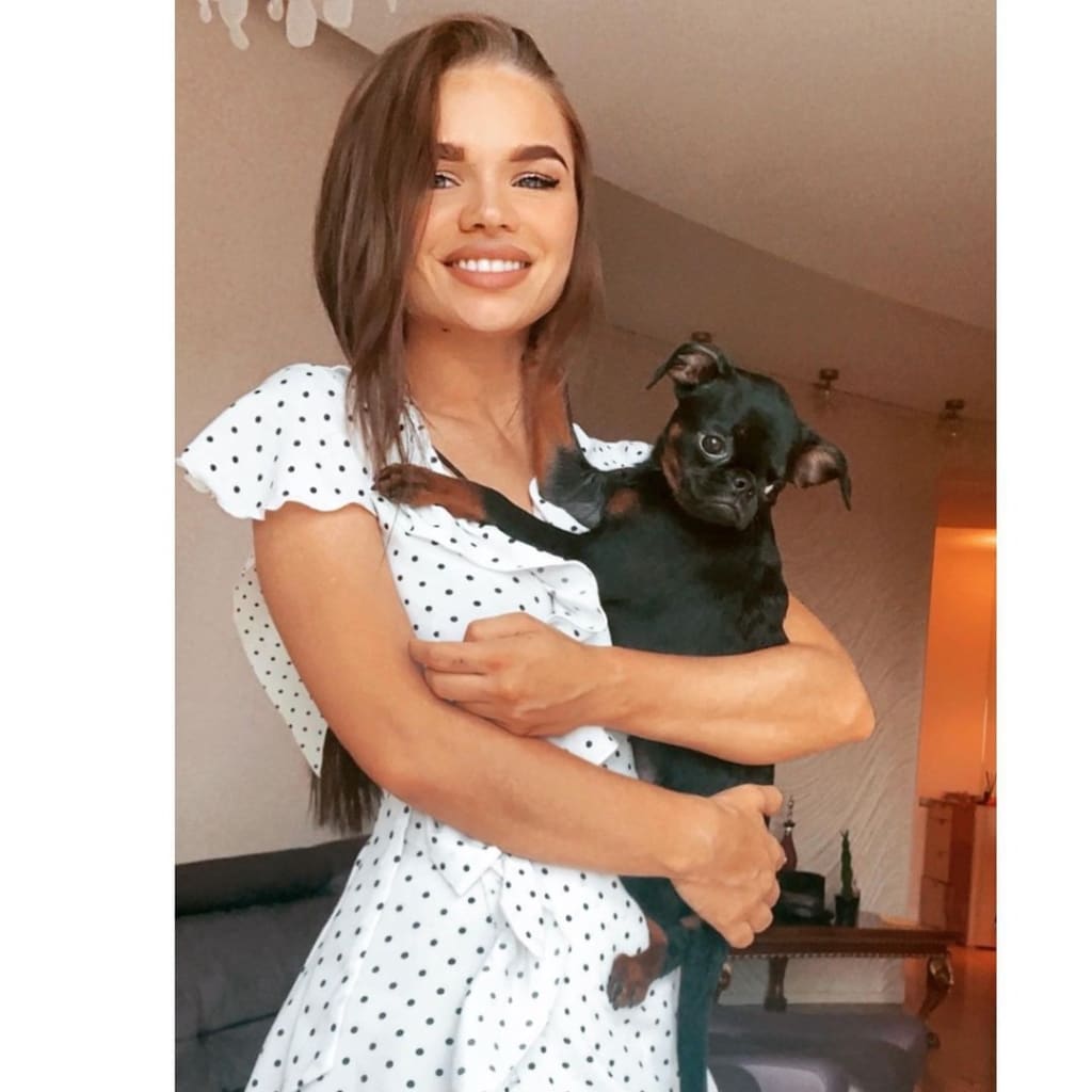 Zoya stands indoors holding a small black dog, wearing a white polka-dot dress with a ruffled neckline, near a sofa and table.