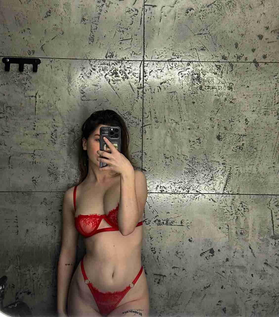 A woman in red lace lingerie stands against a gray wall, holding a smartphone in front of her face.