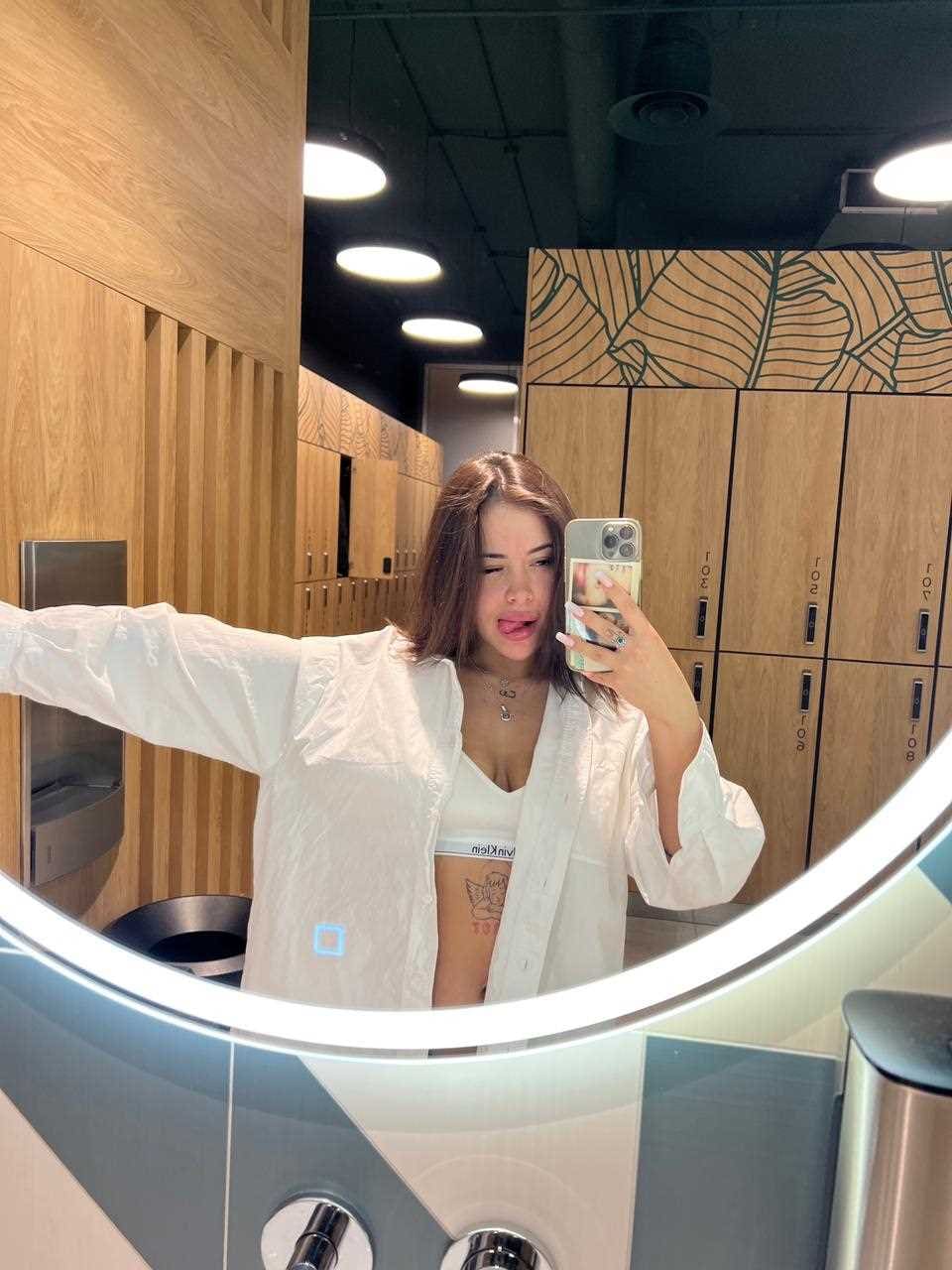 A woman in an open white shirt over a sports bra takes a locker room selfie, revealing a midriff tattoo and wooden lockers.