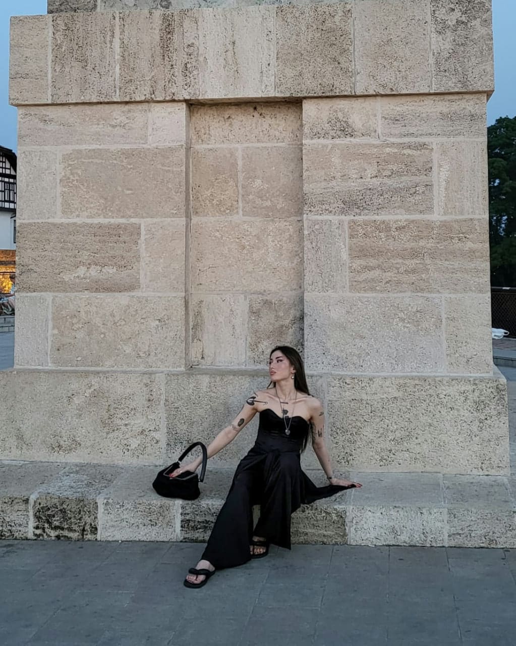 A woman sits on a stone ledge before a stone wall, wearing a black strapless top and wide-leg trousers, holding a black handbag on her lap.