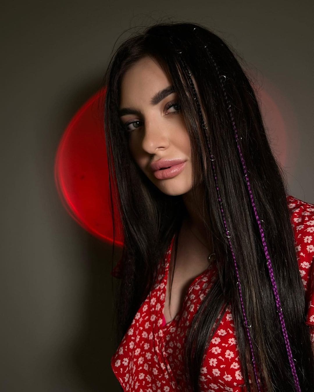 Chloe with long dark hair and purple braids wears a red floral V-neck top against a dark background with a red circular light.