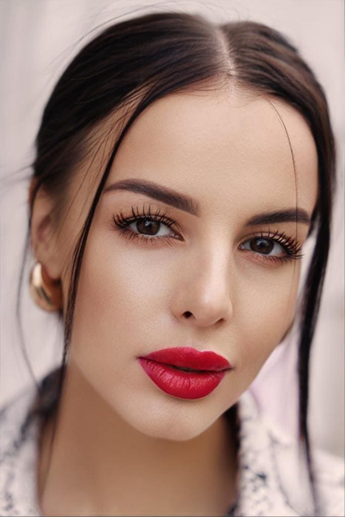 Close-up portrait of a woman with dark hair, red lipstick, and gold hoop earrings against a plain light background.
