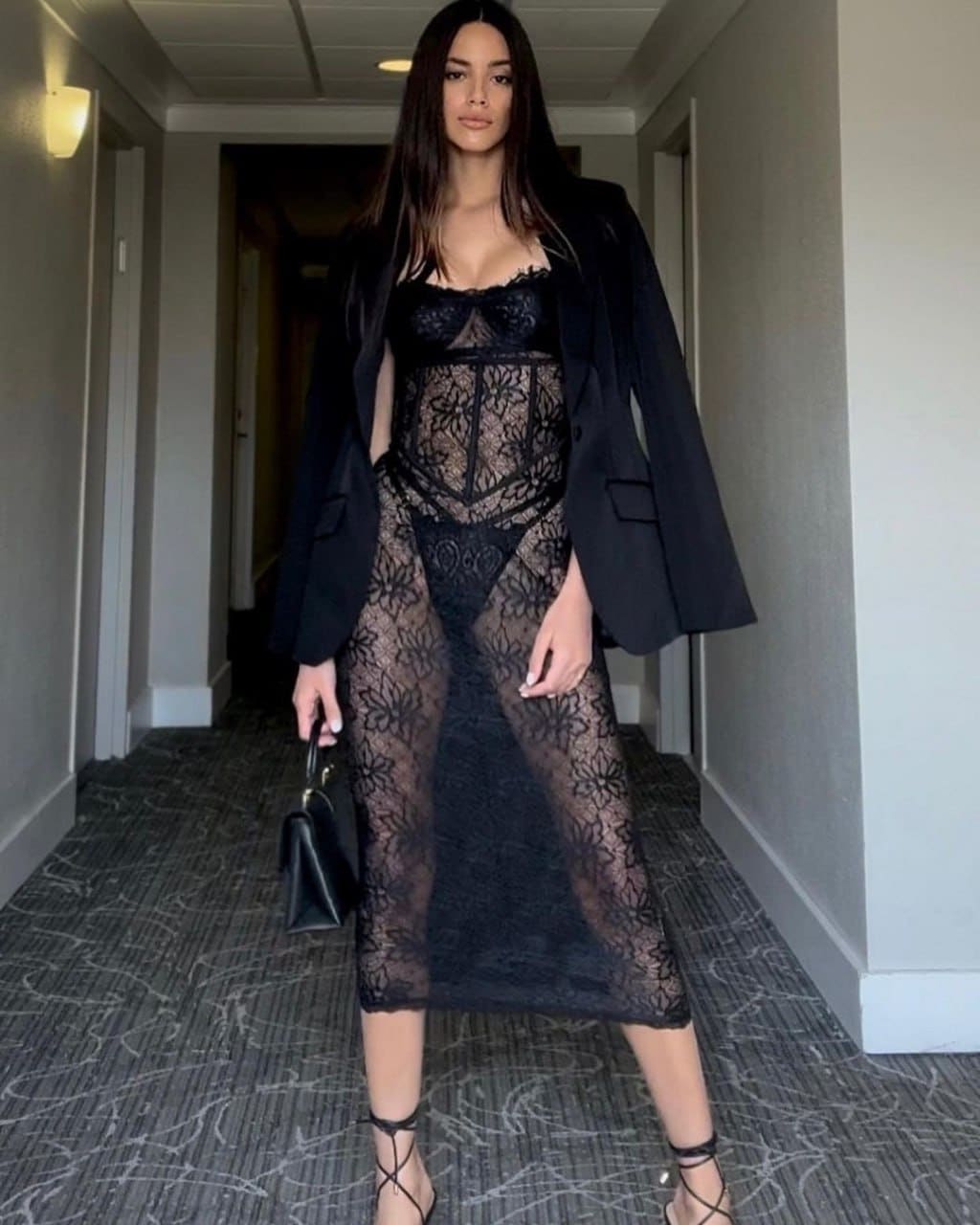 Woman in a sheer black lace midi dress and open blazer stands in a carpeted hallway, holding a small black handbag.