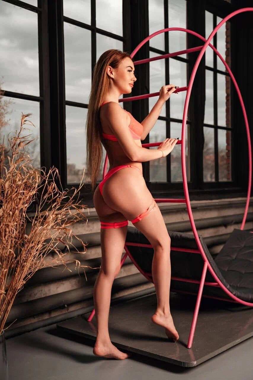 A woman in a red bikini with thigh straps stands barefoot on a dark platform near a pink frame and large windows with dried plants.