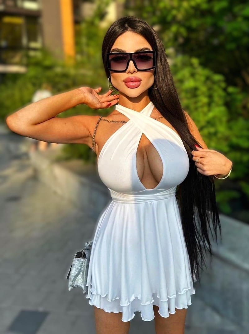 A woman in a white halter mini dress and sunglasses poses outdoors, one hand near her face, the other in her hair.