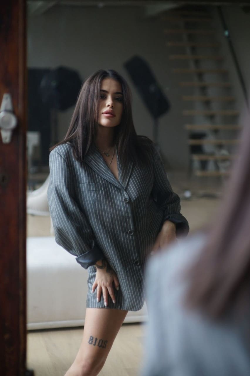 A woman in a pinstriped shirt dress with rolled sleeves stands near a staircase, her hand resting on her thigh.