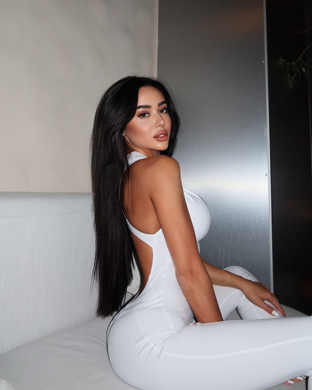 A woman sits on a white couch in a white one-piece outfit with an open back, looking over her shoulder near a metallic wall.