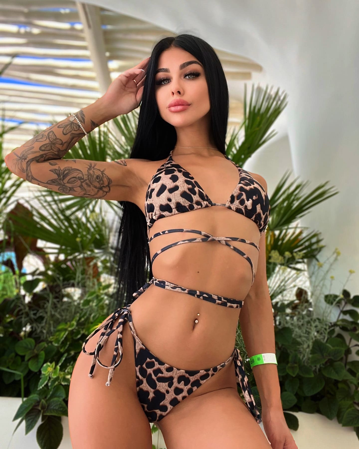 A woman with floral arm tattoos stands in a leopard-print strappy bikini, one hand to her head, before green plants under a white slatted roof.