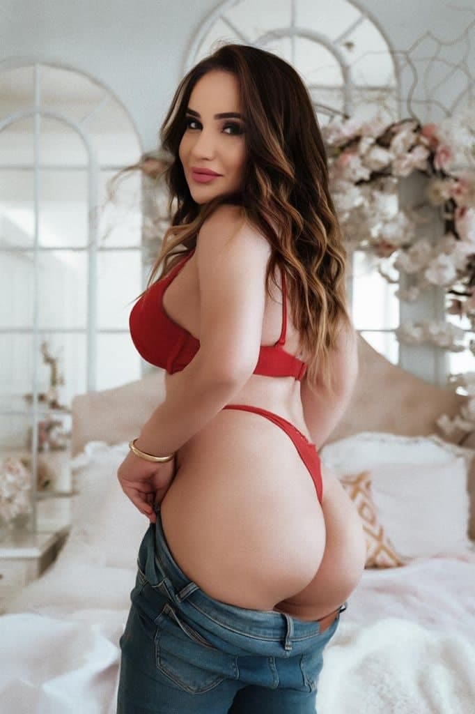 A woman stands in a bedroom wearing a red bra and thong with jeans pulled down to hips, posing with her back partially turned.