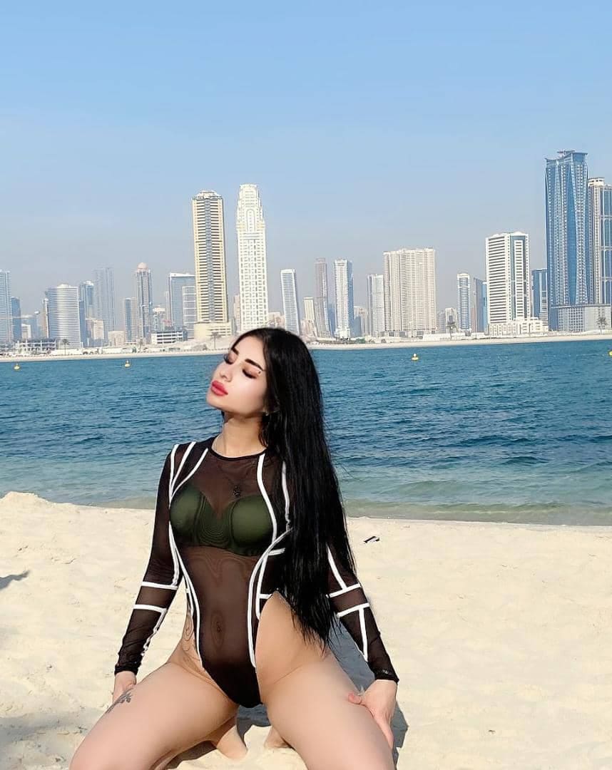 A woman kneels on sand in a sheer black bodysuit over a green bikini, with a city skyline and water visible in the background.