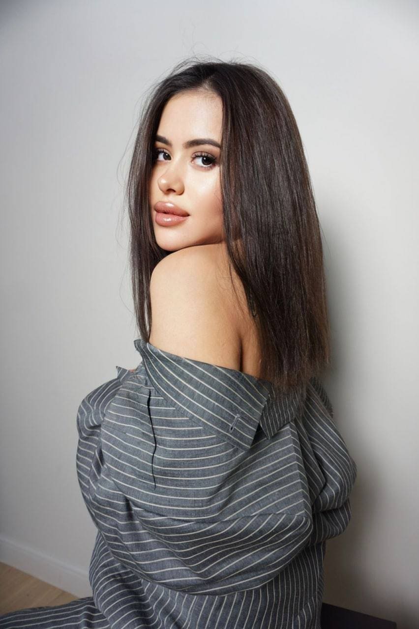 Masha sits in a grey pinstriped shirt worn off the shoulder, looking over her bare shoulder against a plain light wall.
