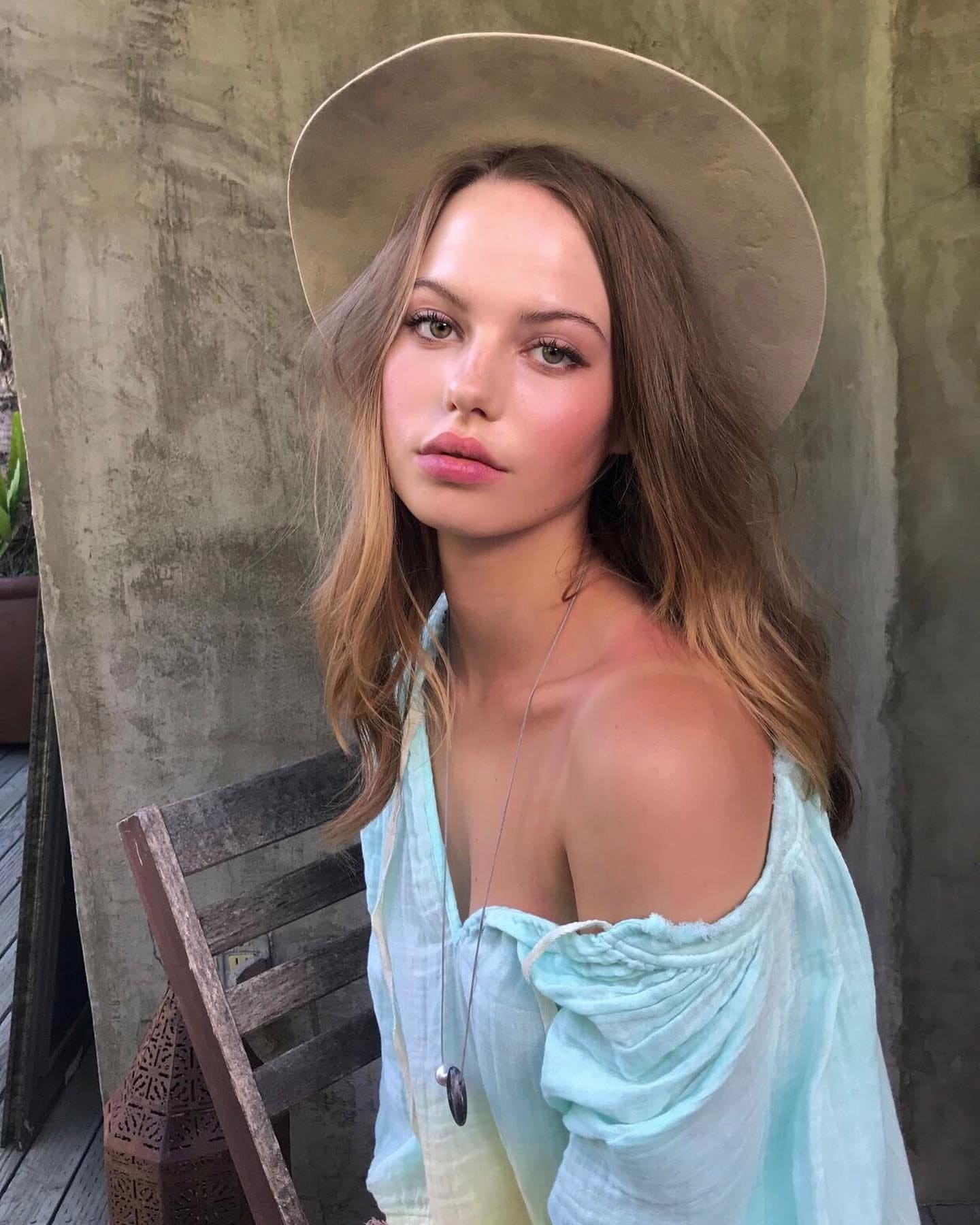 A woman in a beige hat and blue off-shoulder blouse sits on a wooden chair against a concrete wall.