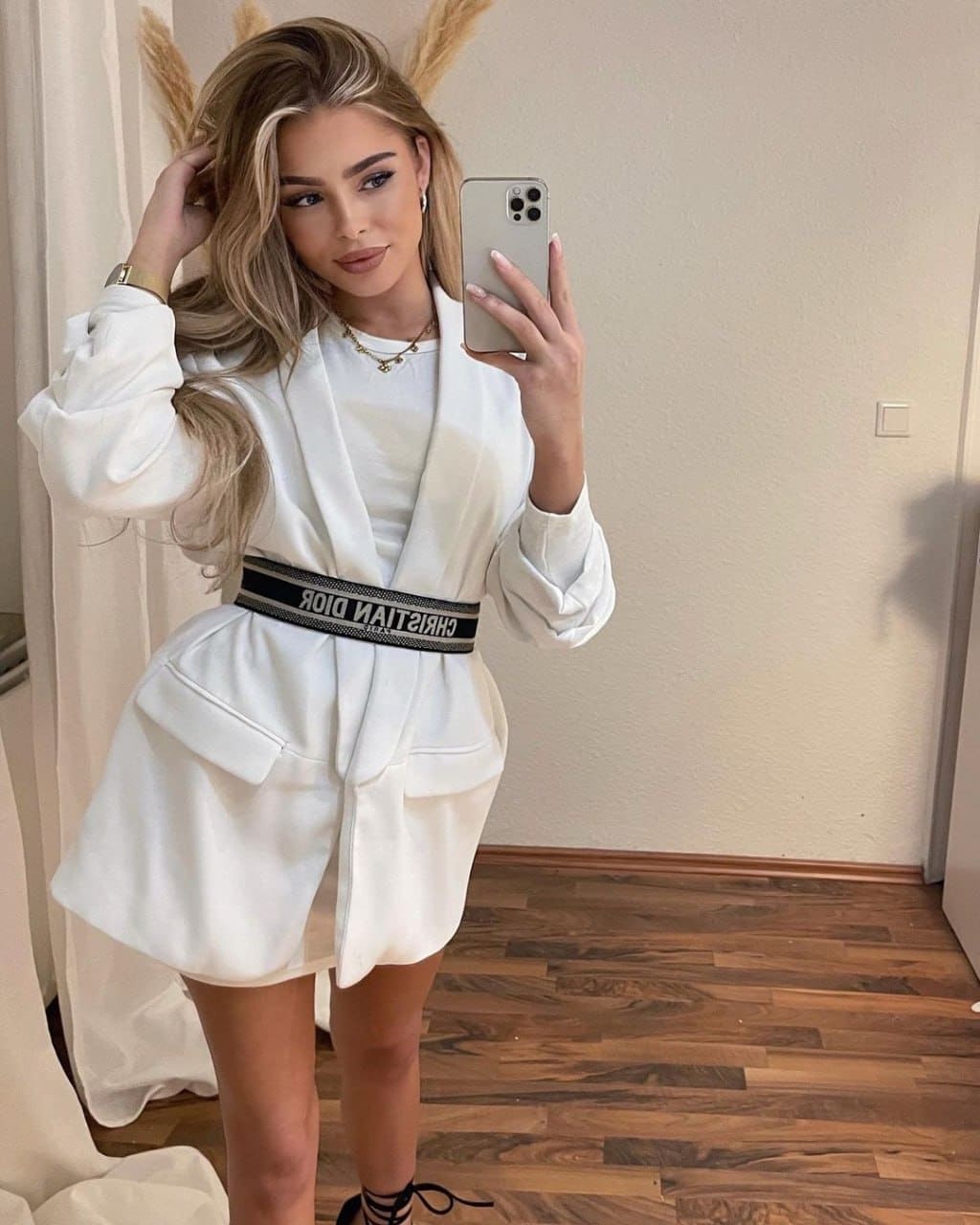 A woman stands on wooden floors wearing a white blazer dress with a black belt, touching her hair while holding a phone.