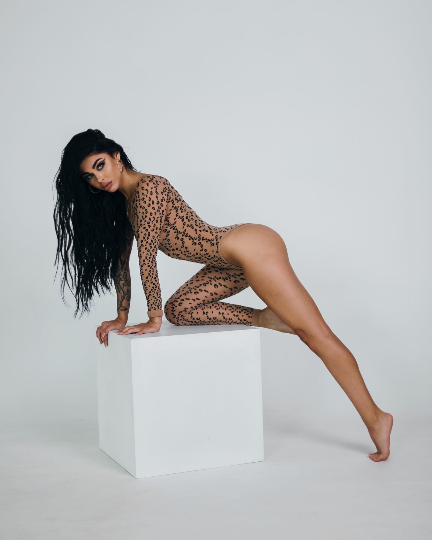 A tattooed woman kneels on a white cube wearing a sheer long-sleeved leopard-print bodysuit, set against a plain light-gray studio background.
