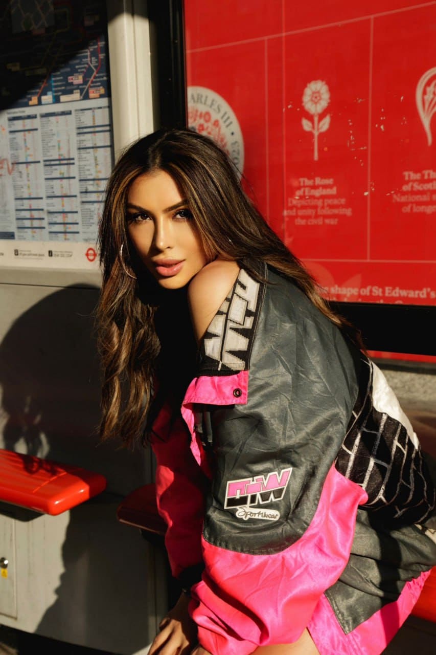 A woman leans against a red subway wall wearing a black and pink jacket with hoop earrings.