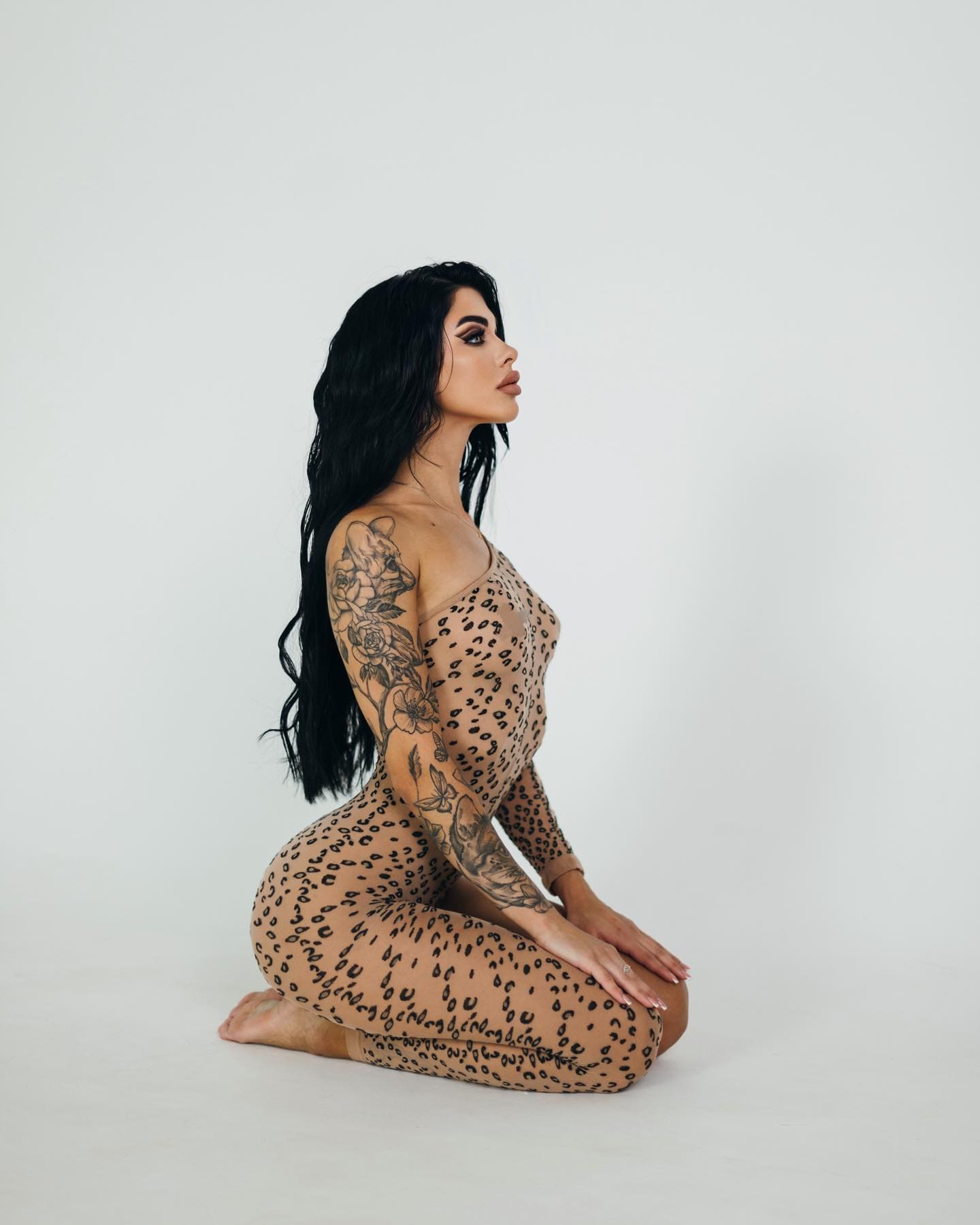 A kneeling woman with floral arm tattoos wears a sheer leopard-print bodysuit over nude fabric on a white studio floor, shown in profile.