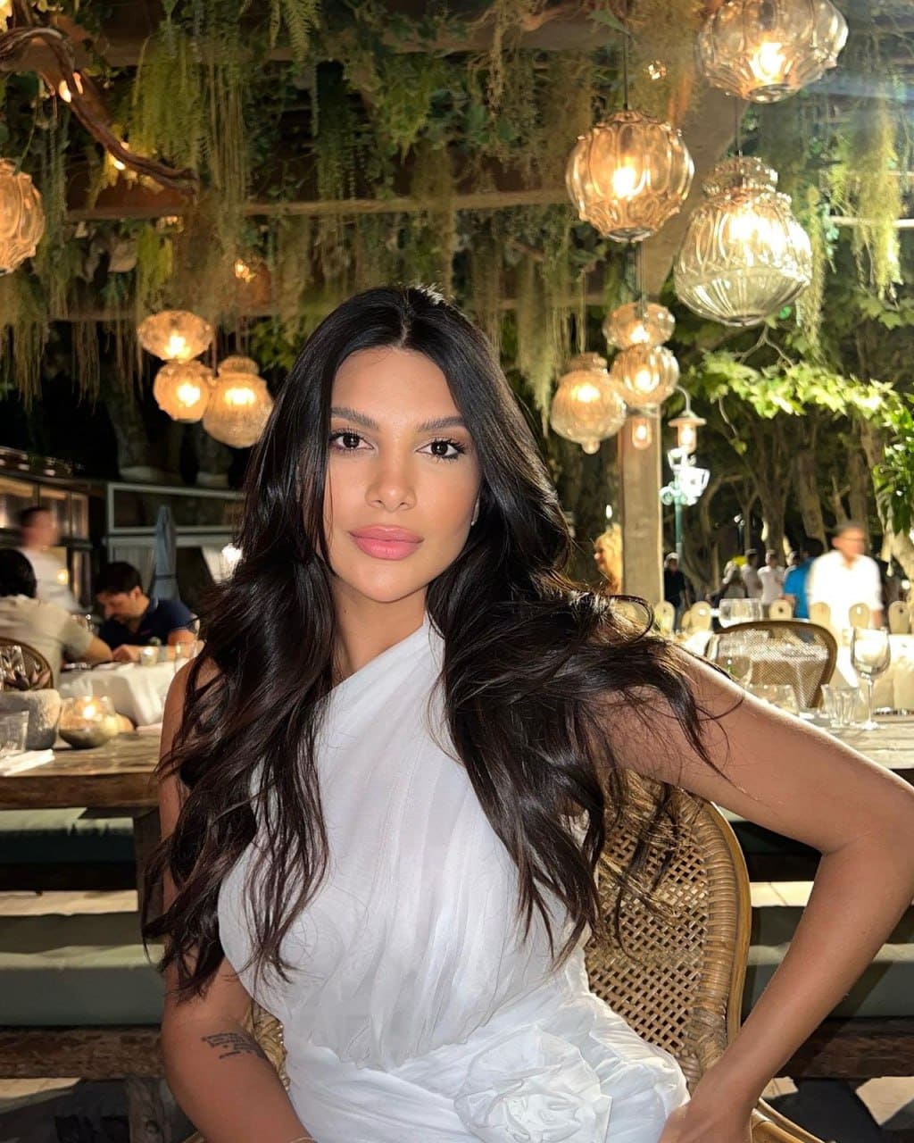 Kath sits in a woven chair at an outdoor restaurant, wearing a white one-shoulder dress, with glass lanterns and greenery behind her.