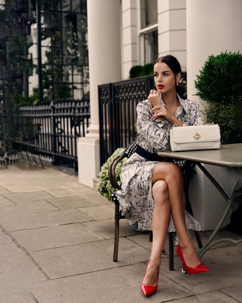 Sitting at an outdoor table, she wears a long-sleeved black-and-white dress with a black belt and red heels, a handbag beside her.