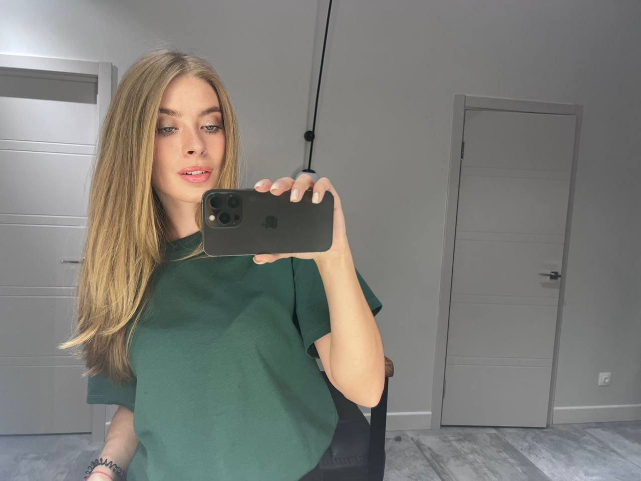 A blonde woman takes a mirror selfie in a loose green t-shirt inside a room with grey walls and closed doors.