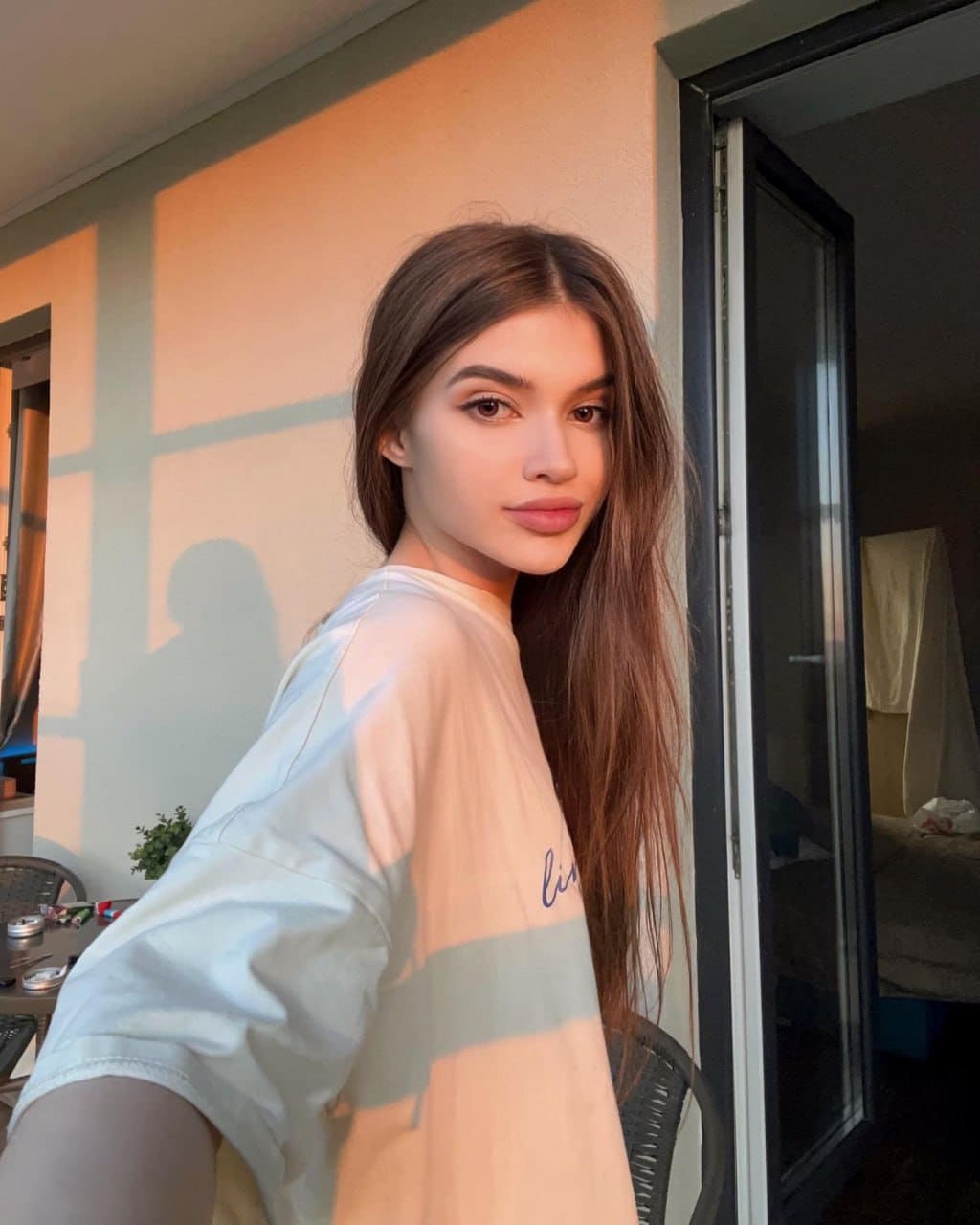 Amelina stands in a sunlit doorway wearing a loose white t-shirt, looking back over her shoulder toward the camera.