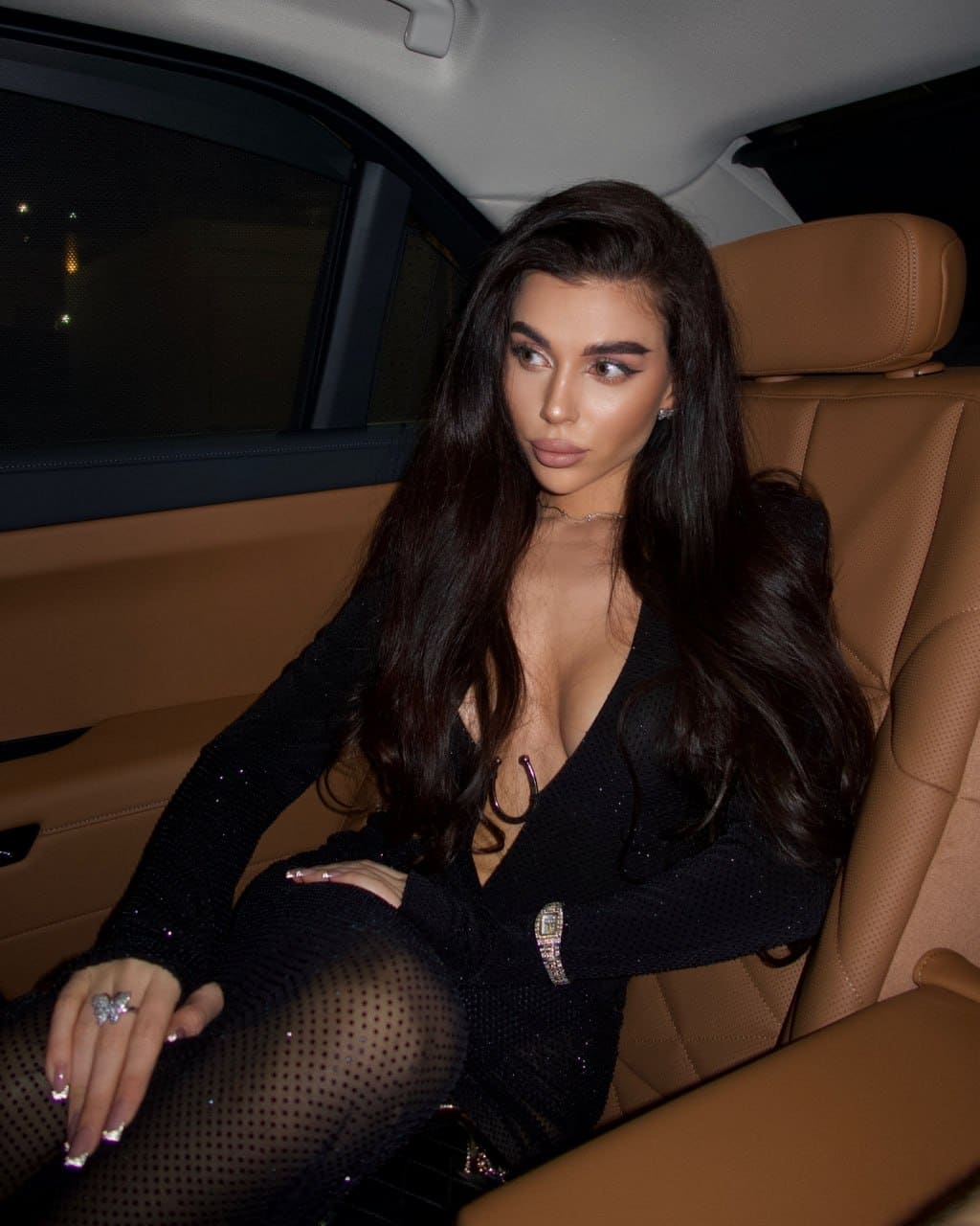 A woman sits in a car with tan leather seats, wearing a black sparkly top and polka-dot tights, her hand resting on her thigh.