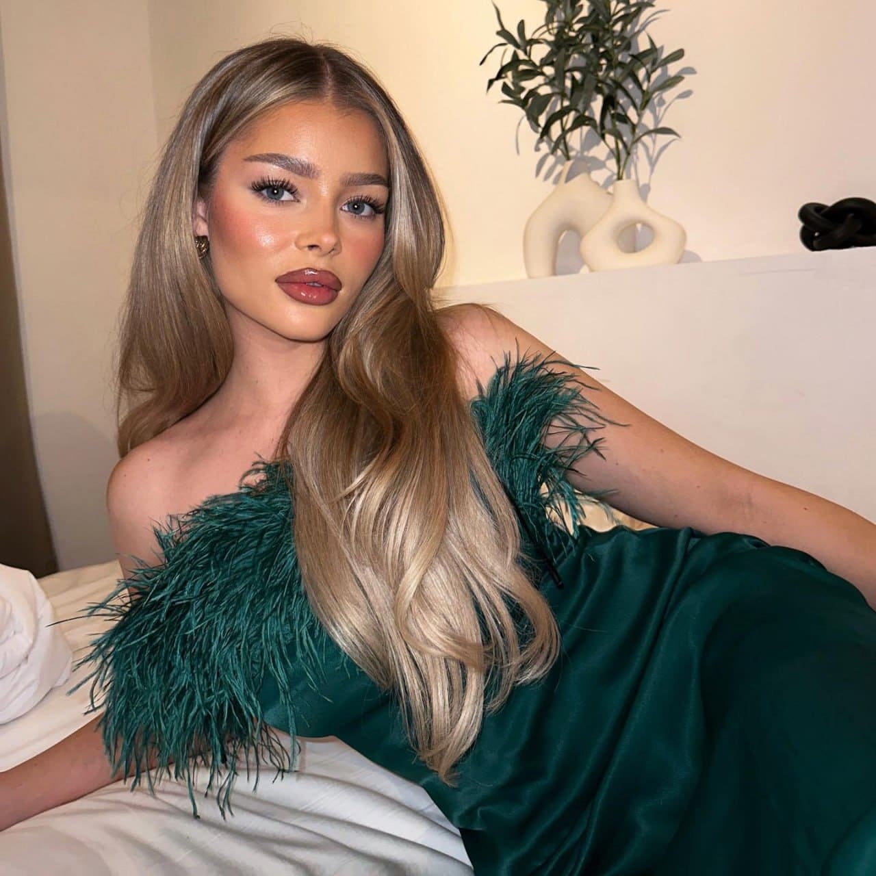 A woman reclines on a white bed in a dark green off-the-shoulder dress with feathered trim, near a potted plant.