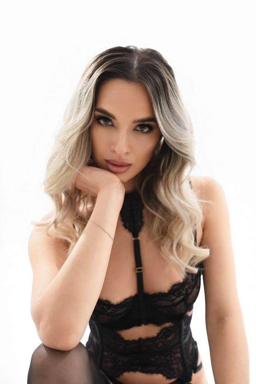 Augusta rests her chin on her hand, wearing a black lace bodysuit and sheer tights against a plain white background.
