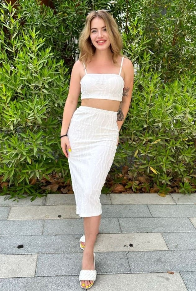 A blonde woman in a white two-piece outfit and slide sandals stands on a paved walkway in front of a green hedge.