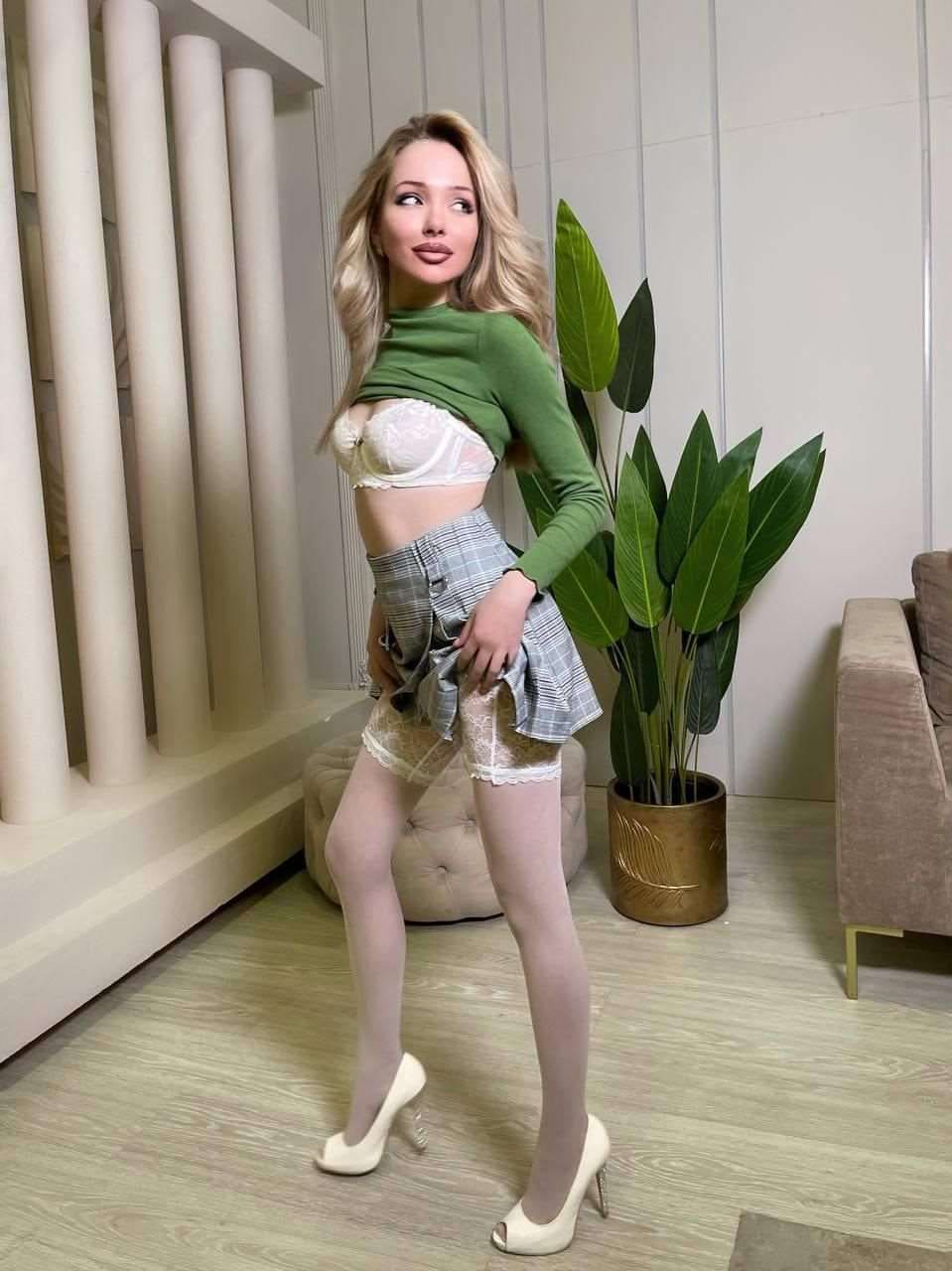 Clara stands in a room with a plant and sofa, wearing a green top, plaid skirt, sheer tights, and white heels.