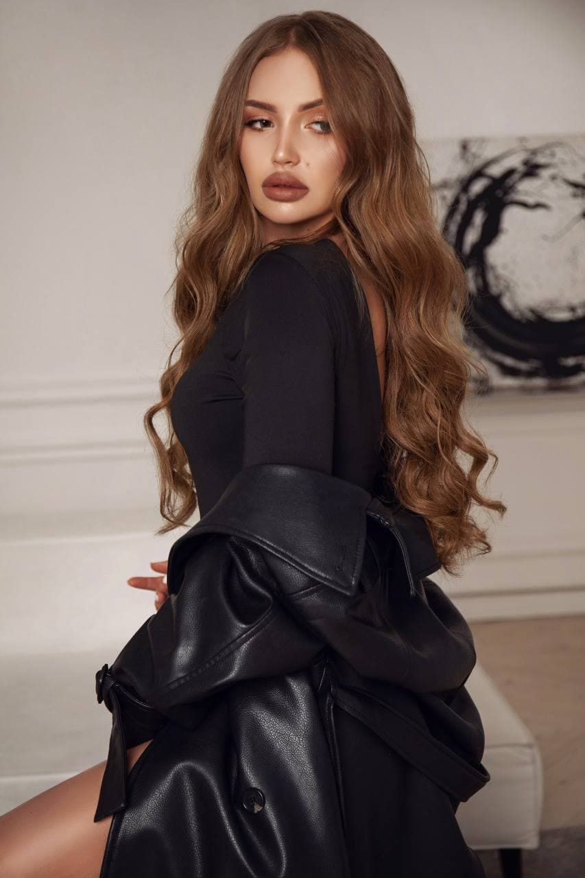 Maria stands in a room with a white wall and abstract painting, wearing a black top and leather coat draped over her arms, showing one bare leg.