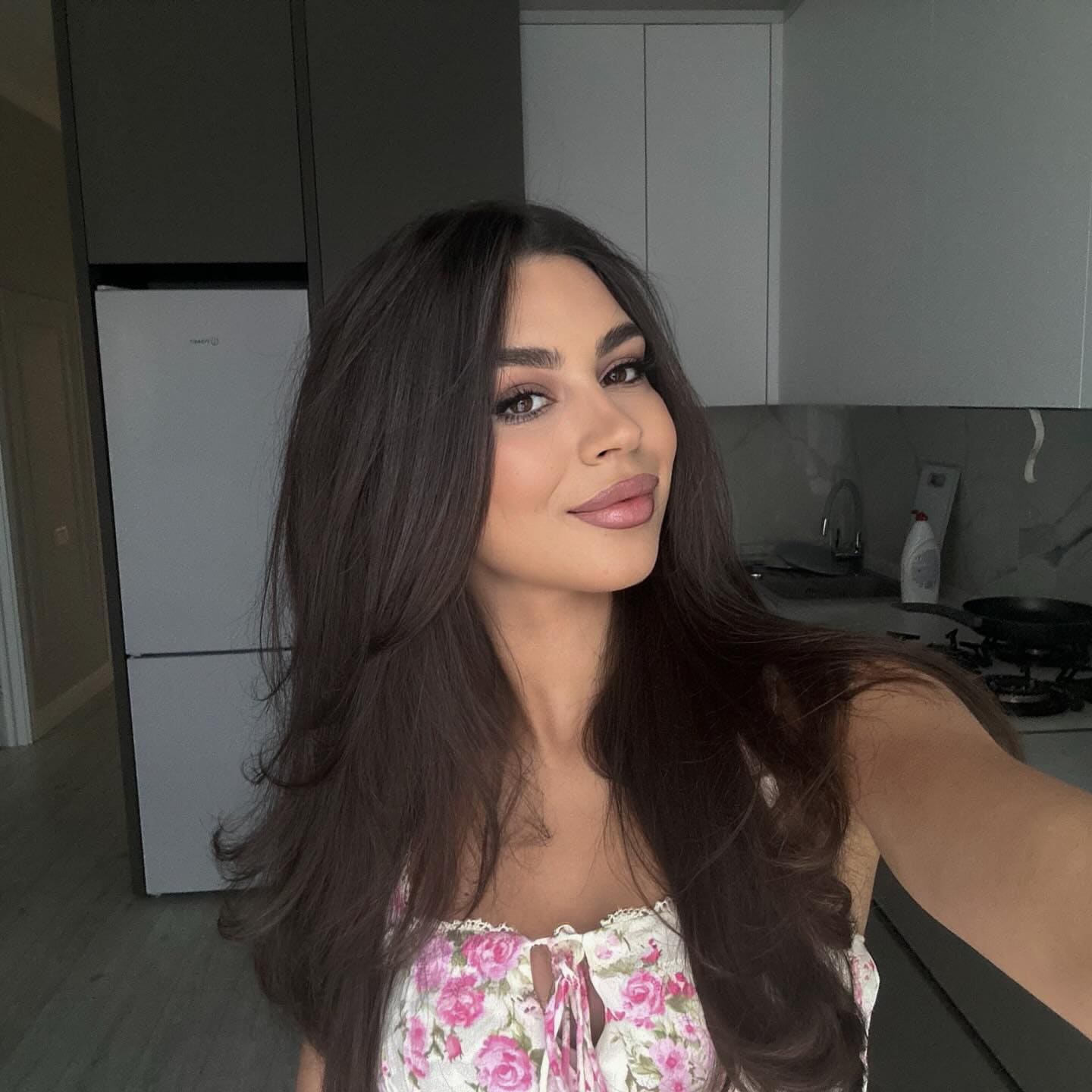 Diana poses in a kitchen wearing a sleeveless floral top with lace trim, with a refrigerator and stove in the background.
