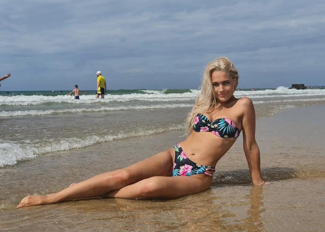 A blonde woman in a floral bikini sits on wet sand at the shoreline, with waves and other beachgoers visible in the background.