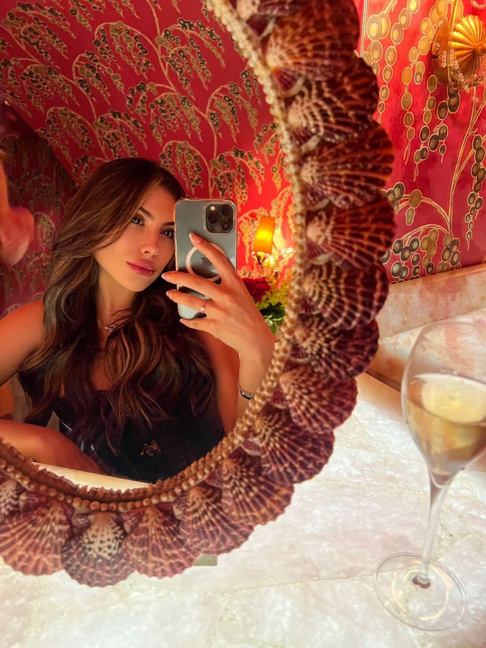 A mirror selfie shows a woman in a dark sleeveless top with a wine glass on the counter behind her.