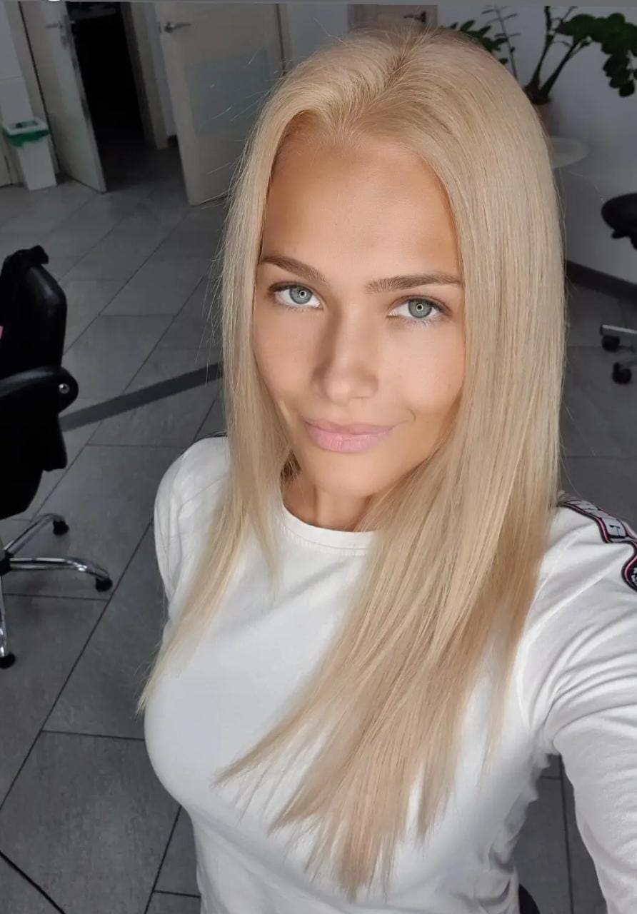 Alexia with long platinum hair wears a white long-sleeved top, taking a selfie in a room with tiled floors and a black office chair.