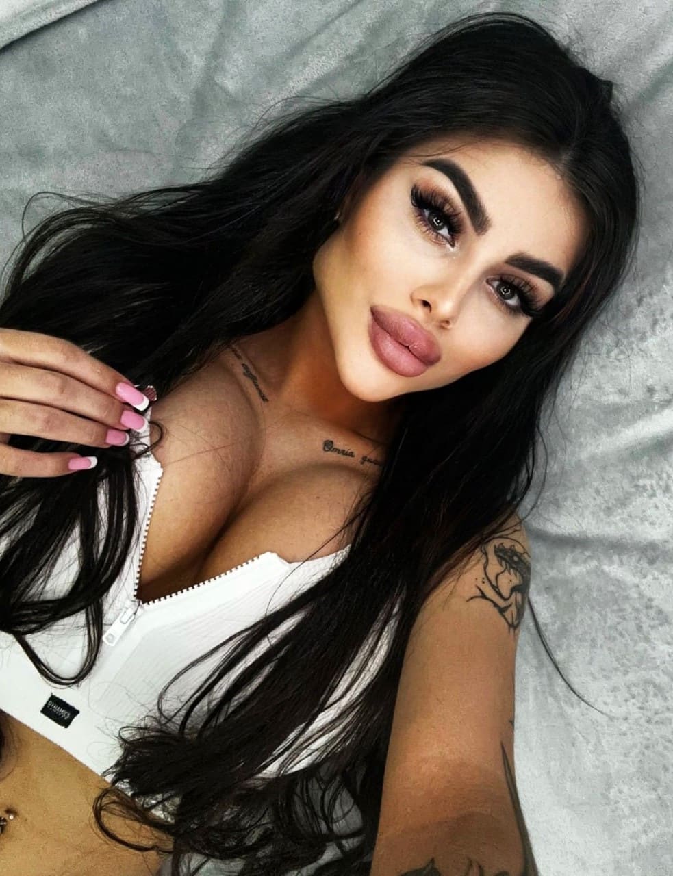 A woman with tattoos lies on grey fabric in a partially unzipped white top, showing pink nail polish.