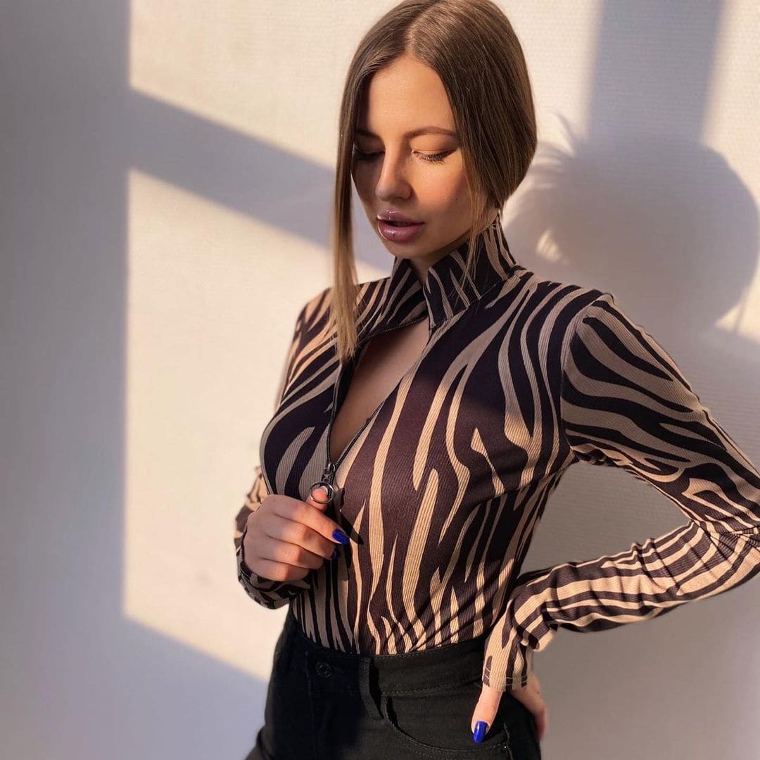 A woman with brown hair wears a zebra-print top and dark pants, holding the zipper while standing against a sunlit wall.