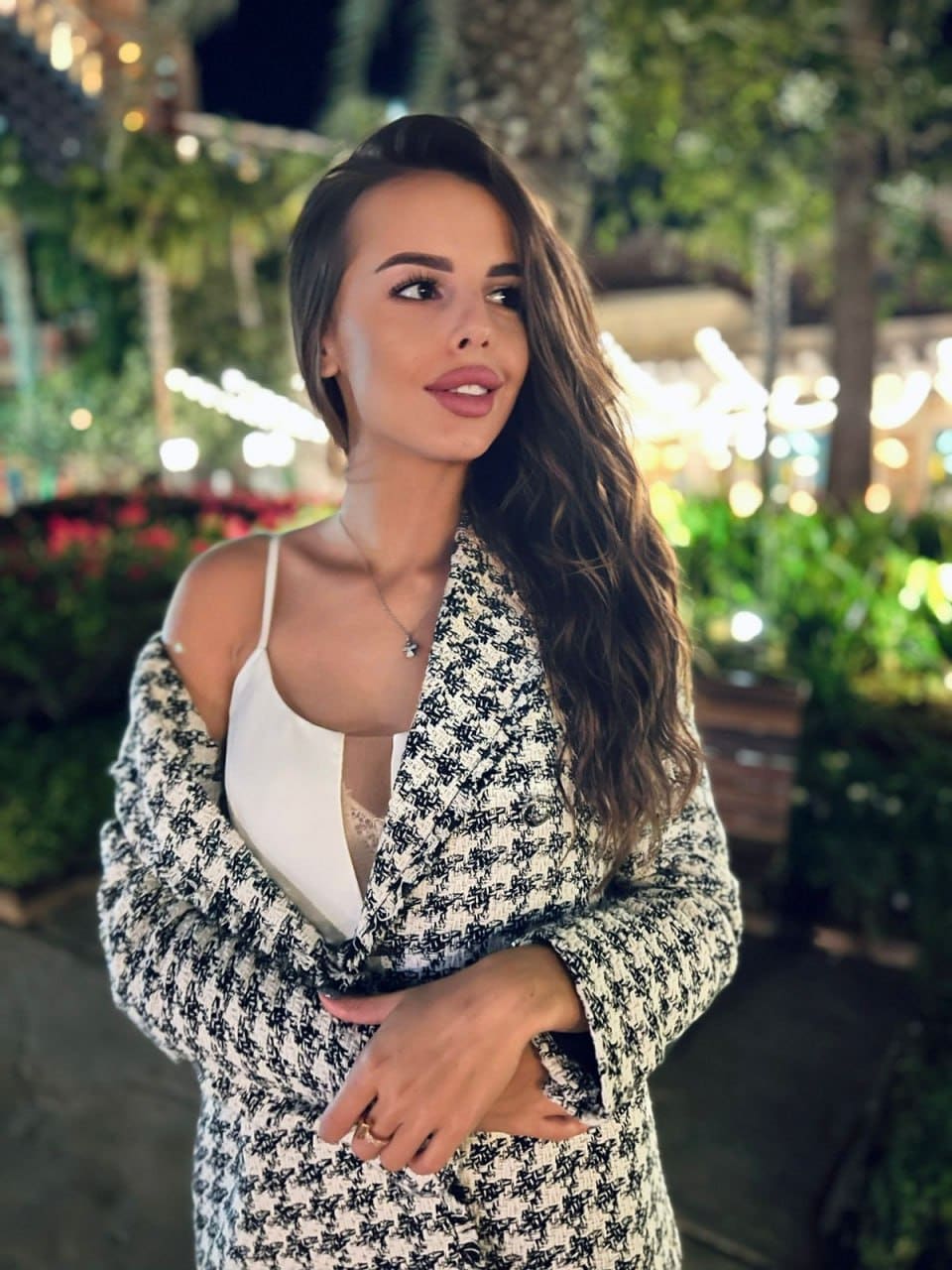 STOP___Mika stands outdoors at night in a black-and-white patterned jacket over a white top, arms crossed, with blurred lights behind.