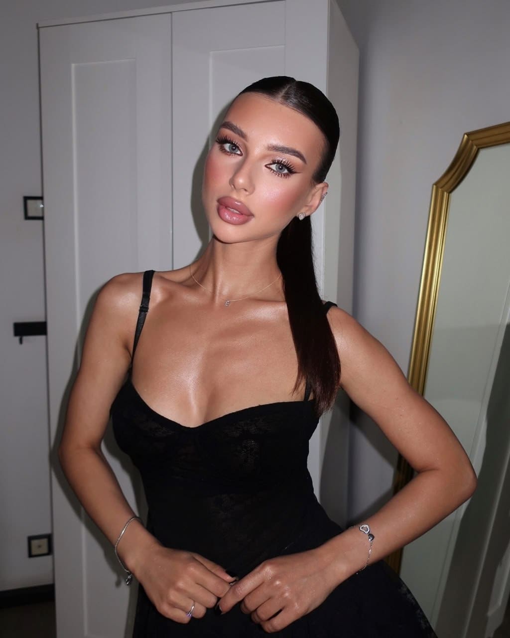 A woman with a ponytail wears a black lace dress with thin straps, standing before a white wardrobe and gold-framed mirror.