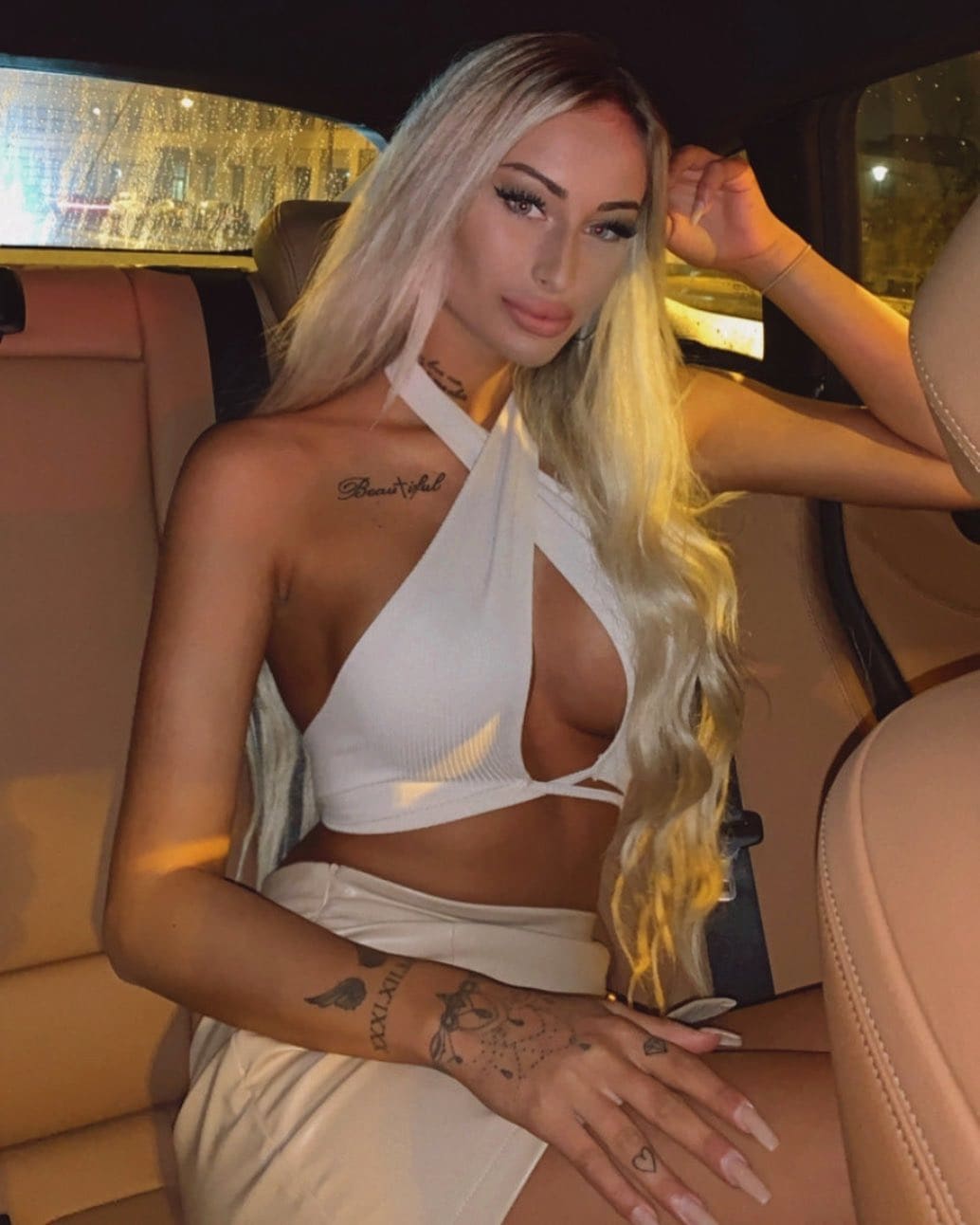 A blonde woman with tattoos sits in a car at night wearing a white halter crop top with side cutout and light skirt.