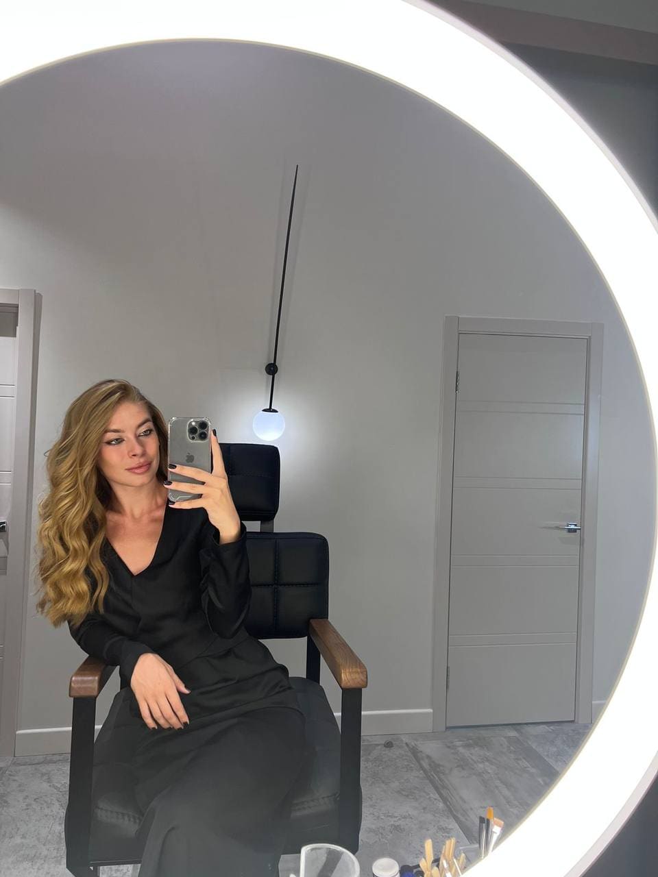 A blonde woman in a black V-neck dress sits in a leather chair, taking a mirror selfie near an illuminated round mirror.