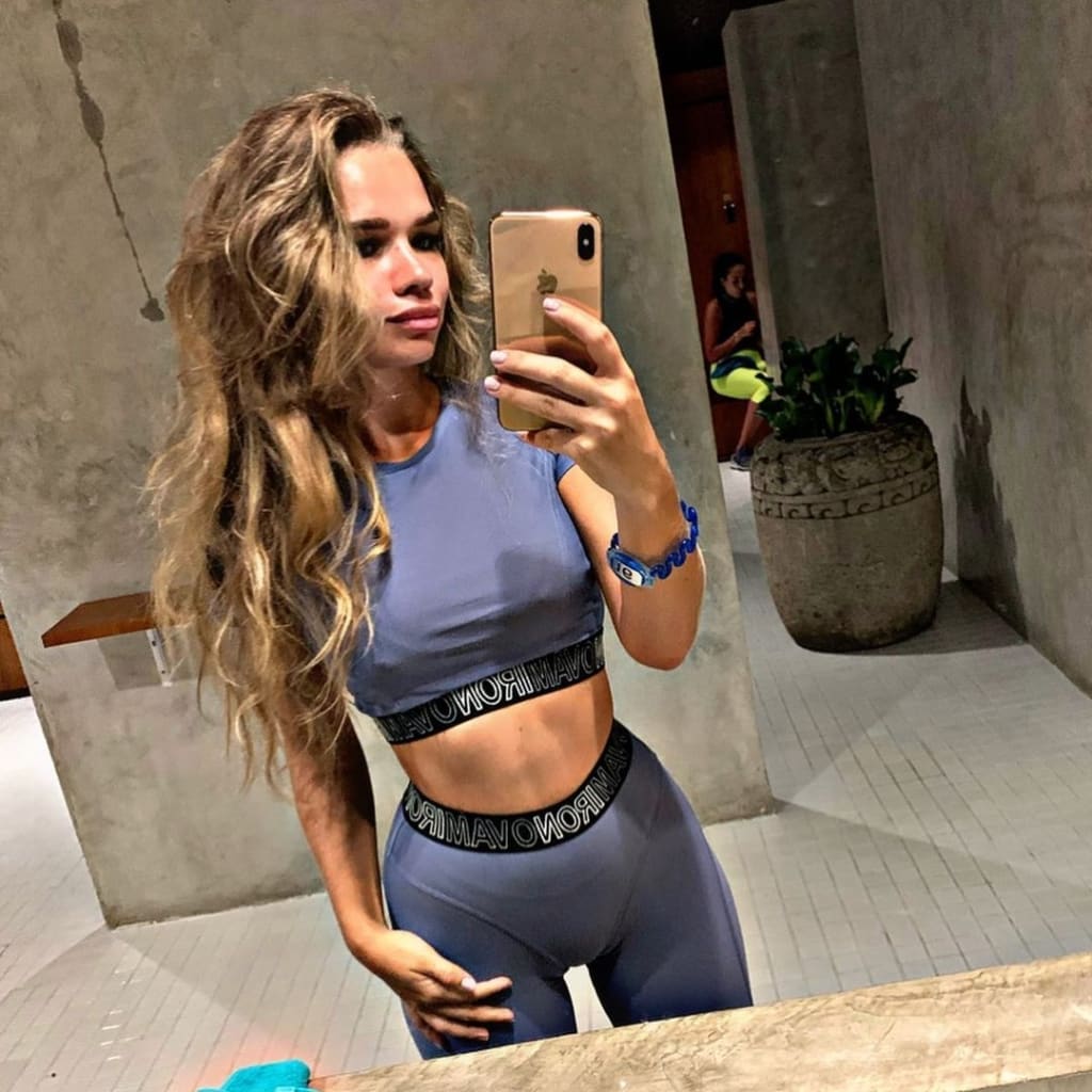 A woman with long wavy blonde hair takes a mirror selfie in a grey crop top and leggings, standing in a concrete room with a plant.
