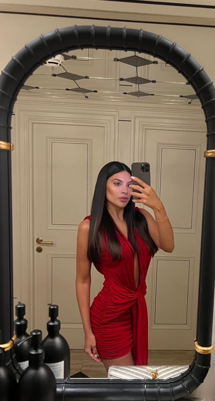 A woman takes a selfie in front of an arched mirror, wearing a red sleeveless dress with a deep V-neck and tied waist, in a paneled room.