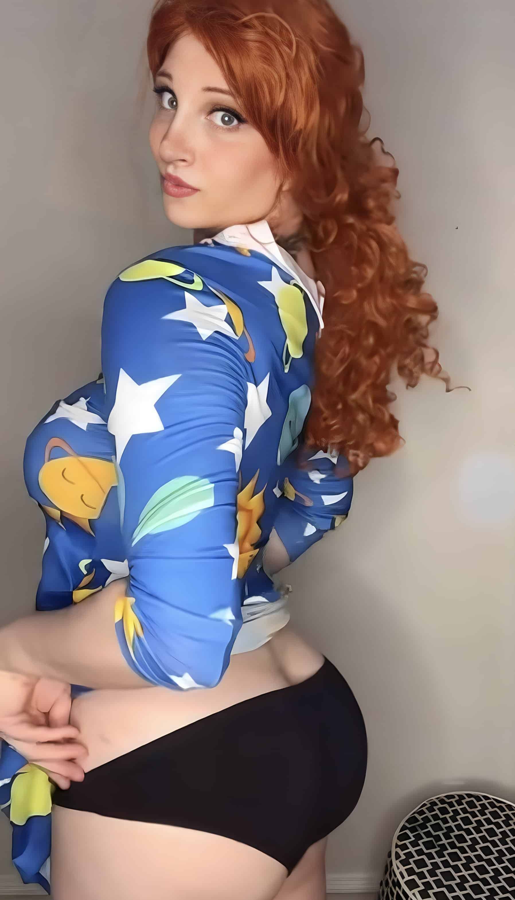 A woman with long curly red hair stands in profile against a plain wall, wearing a blue top with stars and planets and black bikini bottoms, holding the hem.