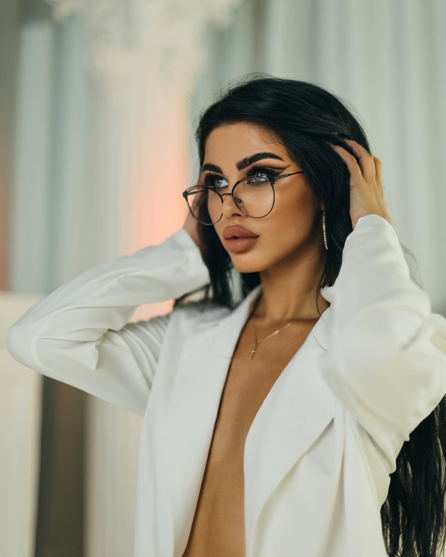 Chidem with long dark hair and round glasses, hands raised to her head, wearing an open white blazer in a lit interior with pale columns.