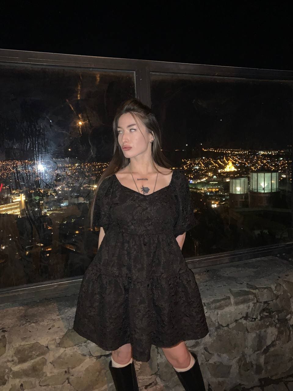 A woman stands on a night terrace in a short black lace dress and knee-high boots, with a lit cityscape visible through a large window.