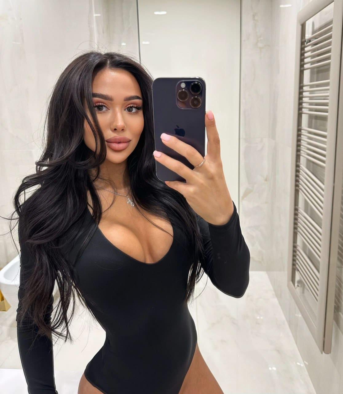 A bathroom mirror selfie features a woman in a black long-sleeved bodysuit with a deep scoop neck, holding a smartphone.