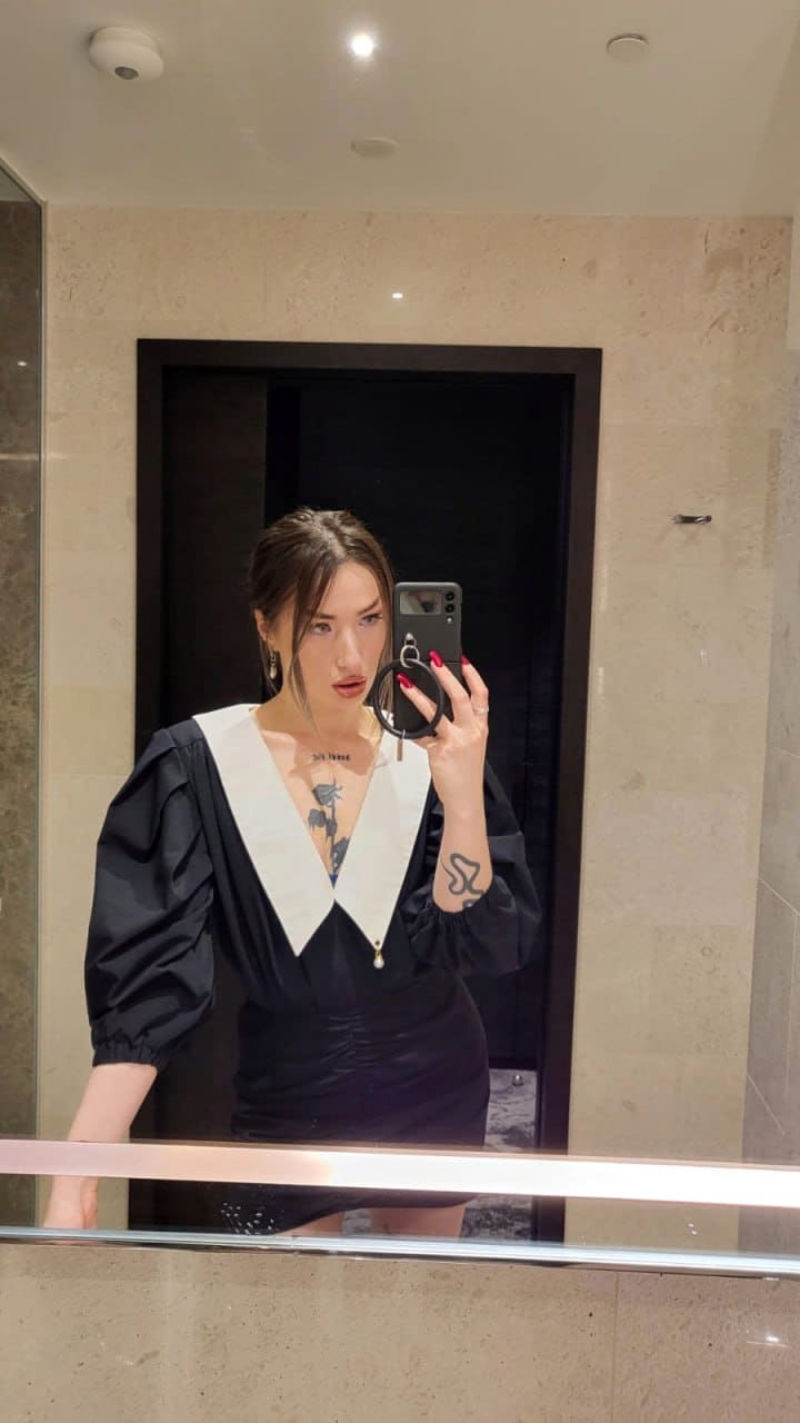 A woman takes a mirror selfie in a black dress with puffed sleeves and a white V-neck collar, revealing a chest tattoo in a beige stone room.