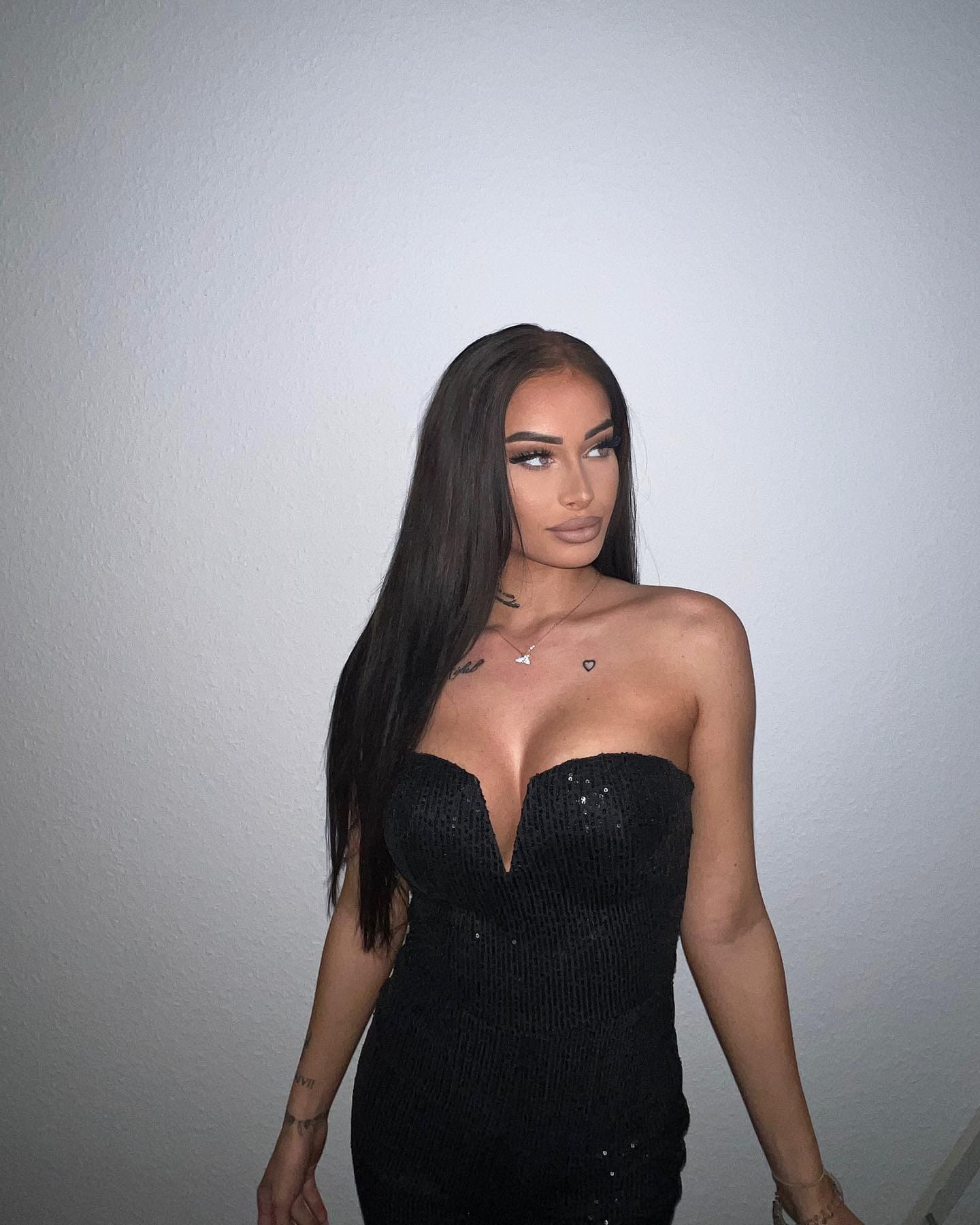 A woman stands with arms relaxed in a black strapless sequined dress with a deep V-neckline and thin necklace against a light wall.