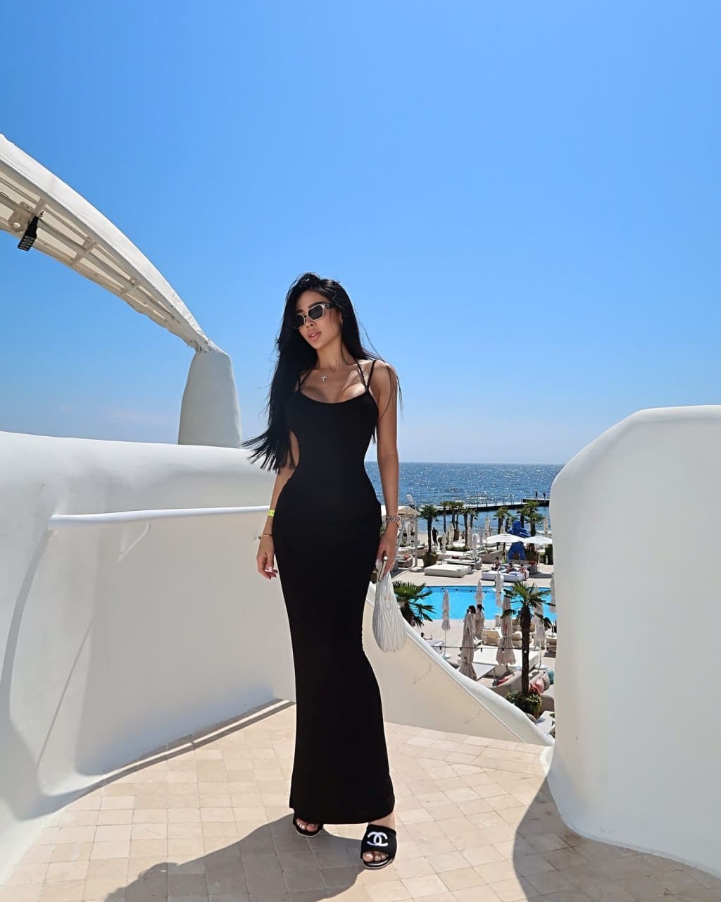 A woman in sunglasses and a long black dress stands on a tiled terrace, with a pool and sea visible under a blue sky.